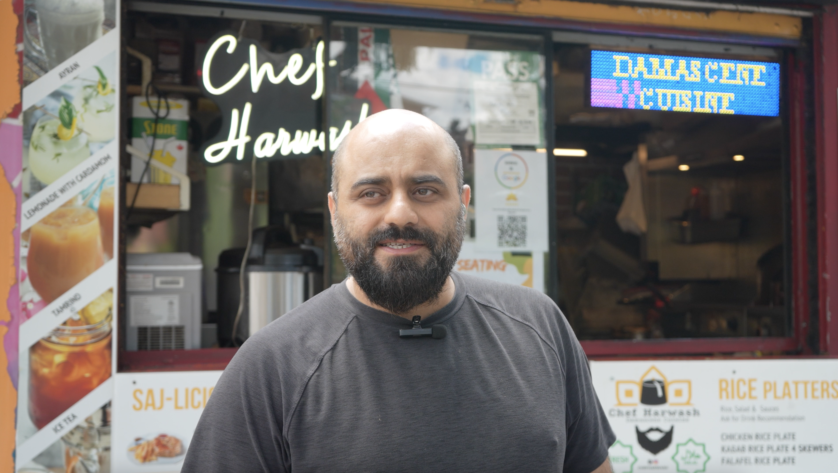 A middle aged man wearing a black T-shirt stands in front of a food stall. Behind him is a white neon logo that reads "Chef Harwash"