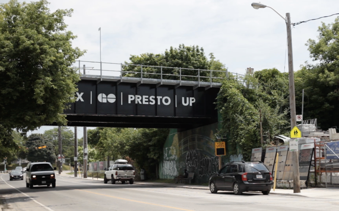 A view of a street lined with trees. Above it is a black bridge that reads GO PRESTO UP