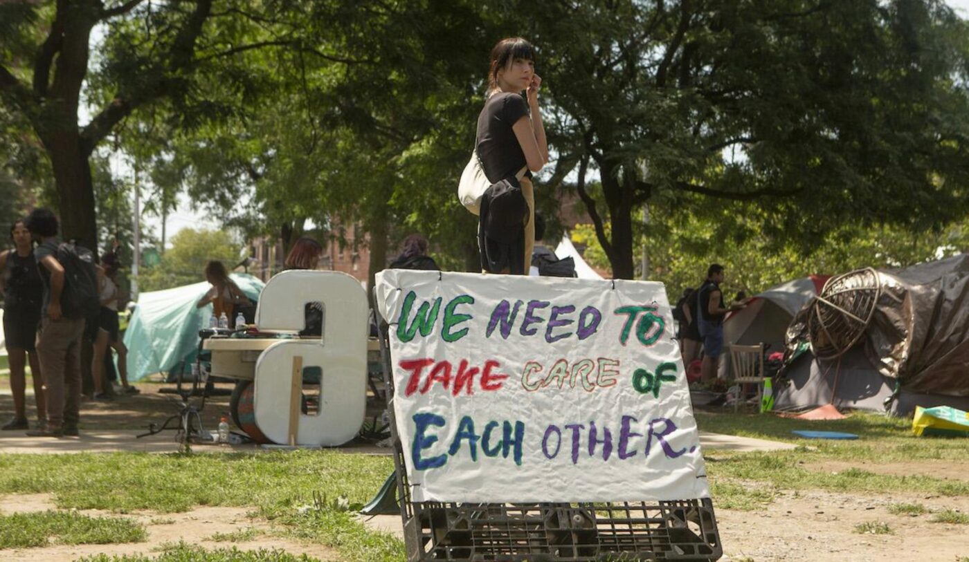 Diana Chan McNally stands in a park, with a sign that reads "We need to take care of each other" in front of her.