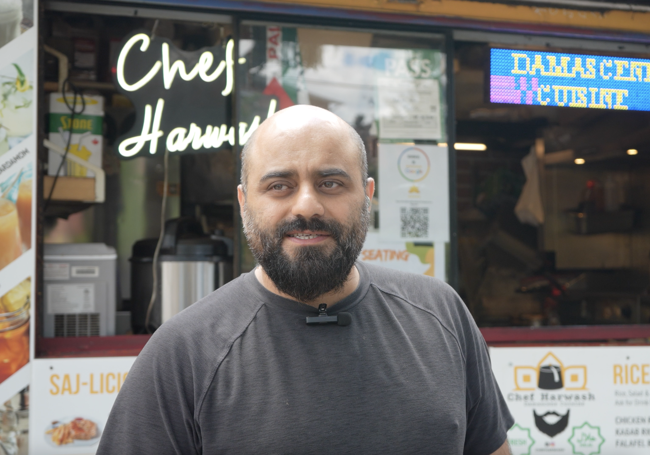 A middle aged man wearing a black T-shirt stands in front of a food stall. Behind him is a white neon logo that reads "Chef Harwash"