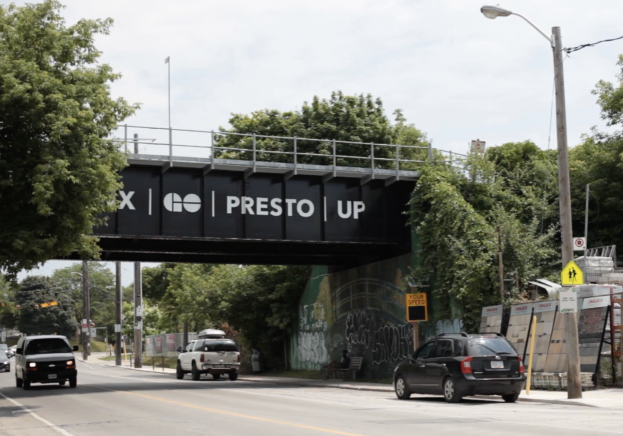 A view of a street lined with trees. Above it is a black bridge that reads GO PRESTO UP