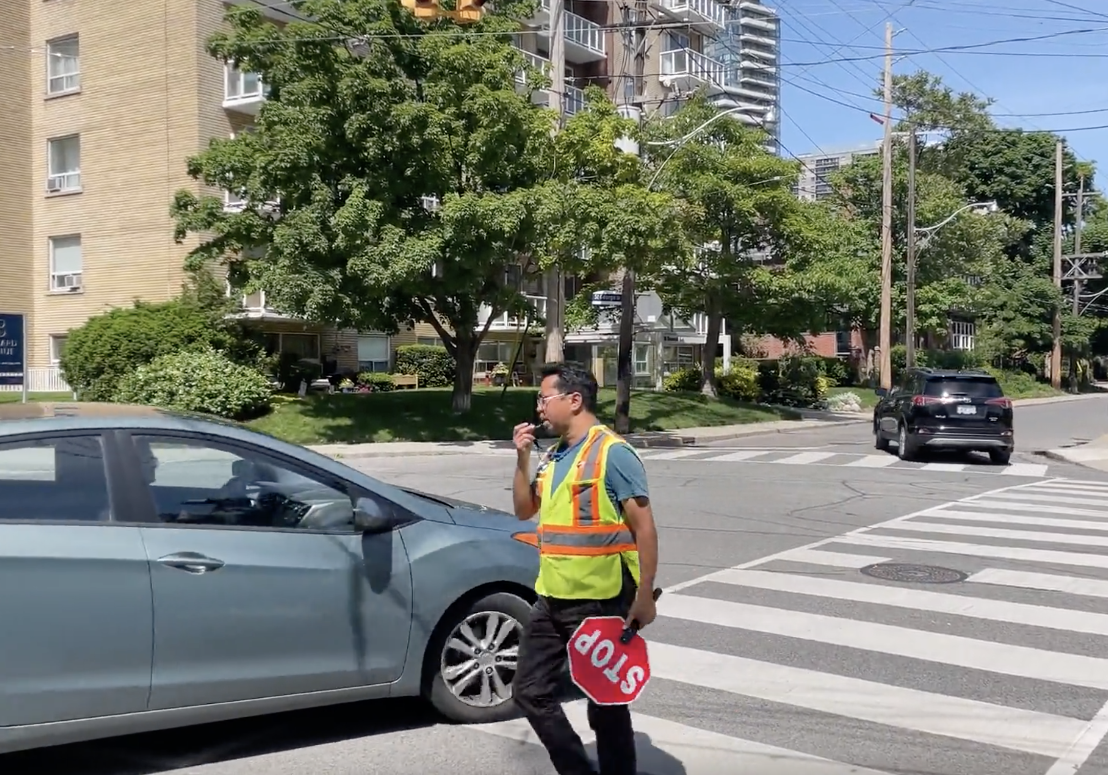 Ulric Singh crosses the street with a stop sign in one hand and a whistle in another.